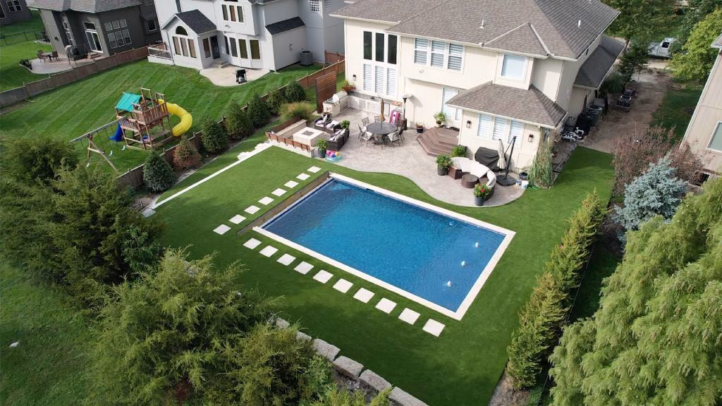 Aerial views of the backside of a house with a beautiful backyard featuring artificial grass, an inground pool, and a stone patio with a fire pit, sitting areas