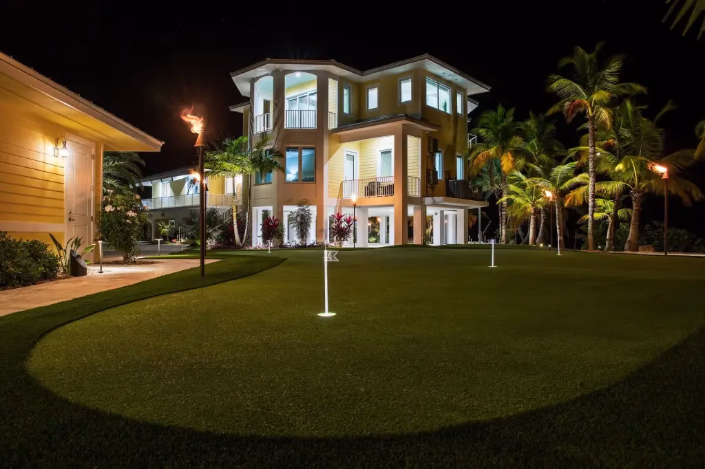 Night photo of a backyard with lighted flags on an artificial putting green with a 3-story yellow house on the background